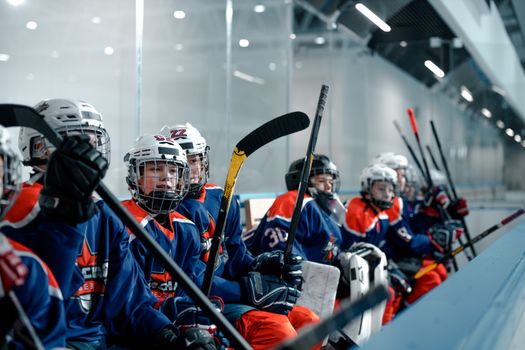 Home A youth ice hockey team sitting on the bench during a game in an ice arena, wearing uniforms and helmets.